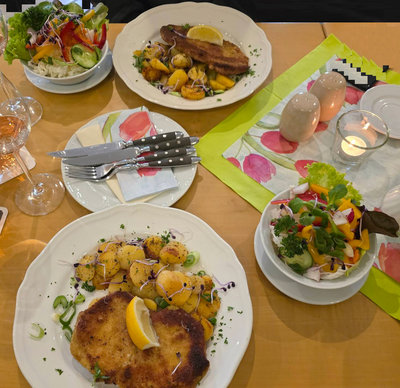 Zwei Teller mit Hauptgerichten, dazu Salate und ein Glas Wein auf einem Tisch.